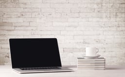 An open laptop on an office desk with flower, coffee, books in front of white brick wall background concept