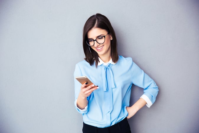 lady wearing a blue shirt standing in front of a gray wall holding her cell phone