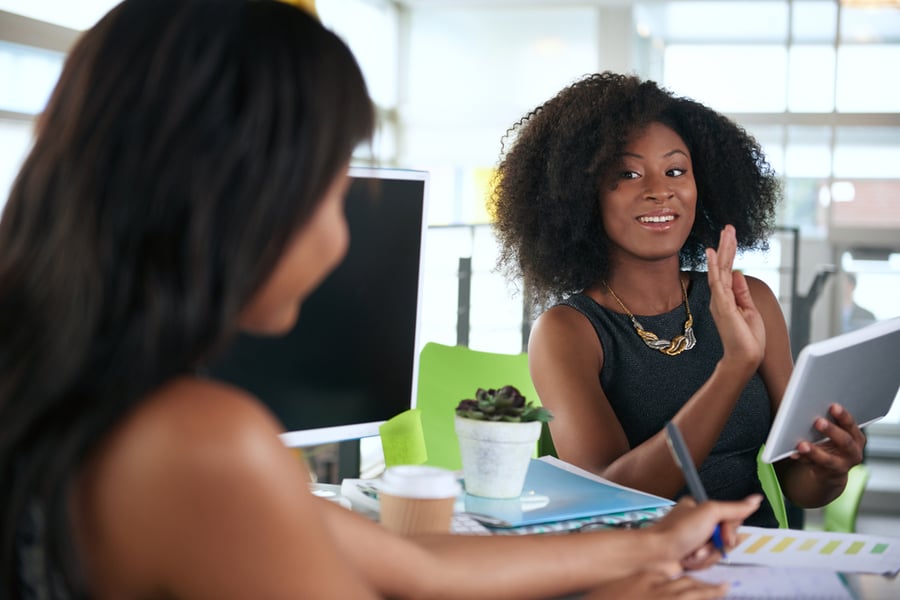 Two ladies talking to one another while sitting at their office desks.