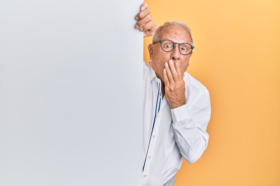 Man peering from behind a white wall with hand over his mouth.