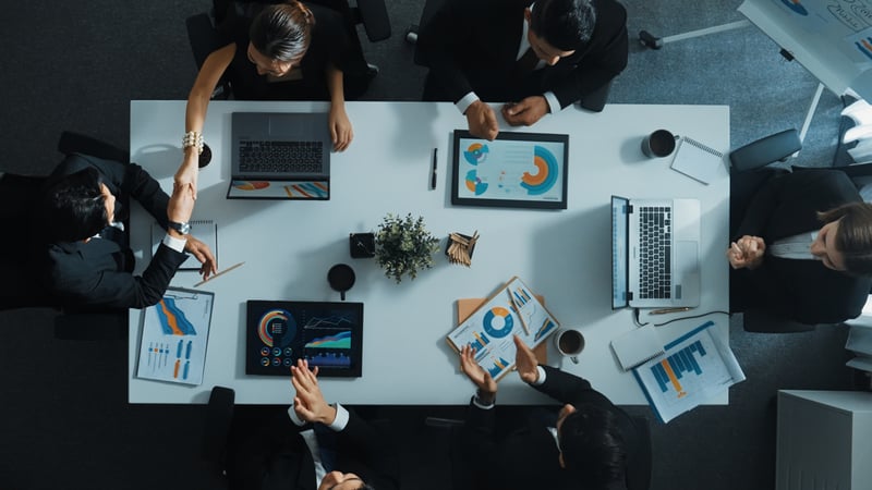 Overhead view of a marketing and sales team collaborating around a conference table with laptops, tablets, charts, and marketing data.